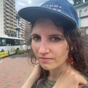 Woman with curly brown hair wearing a baseball cap looks at the camera on a city street.